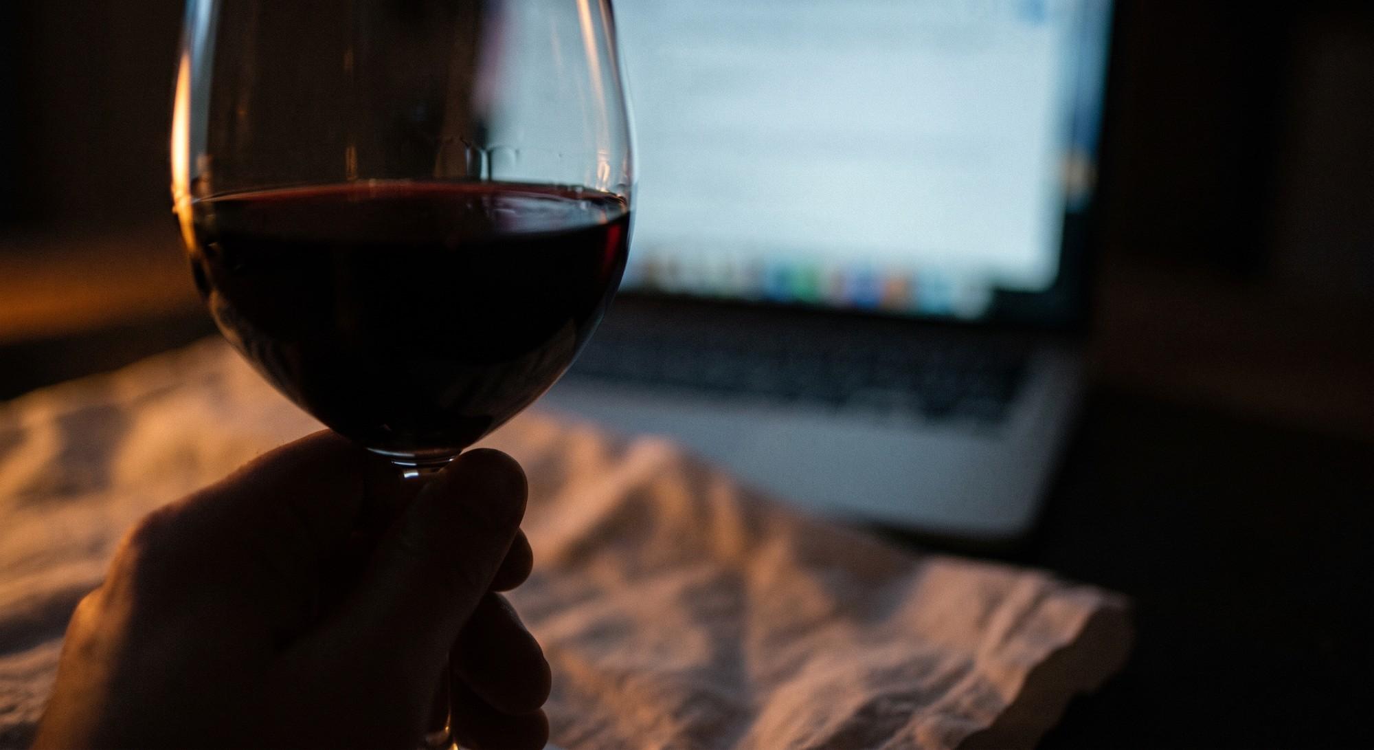 A hand holding a glass of red wine in front of an open laptop in a dim room.